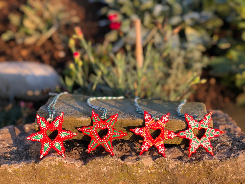 May include: Four red wooden star ornaments with green and white painted details. Each star has a heart-shaped cutout in the center and is attached to a white and green string.