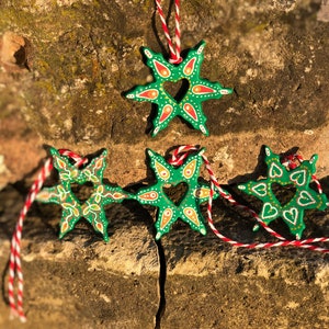 May include: Four green star-shaped ornaments with a heart cut-out in the center. The ornaments are painted with white, red, and yellow details. They are hanging from red and white string.