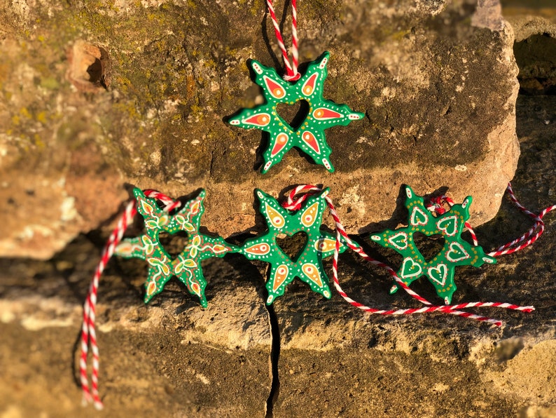 May include: Four green star-shaped ornaments with a heart cut-out in the center. The ornaments are painted with white, red, and yellow details. They are hanging from red and white string.
