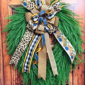 May include: A teardrop-shaped wreath made of green fern-like foliage, adorned with a large multi-layered bow. The bow features ribbons in tan burlap, black and white buffalo check, leopard print, and a pattern of blue and orange pumpkins.
