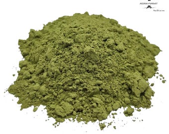 May include: A mound of fine, olive-green powder with a textured, clumpy consistency. The substance is piled in a heap against a plain white background. In the upper right corner, a small black logo features a tree and the text AGORA MARKET.