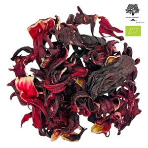 Może przedstawiać: Zbliżenie suszonych kwiatów hibiskusa, prezentujące głęboko czerwone płatki i działki kielicha. Obraz podkreśla naturalną teksturę i wariacje kolorystyczne składnika herbaty. Widoczne jest logo Agora Market i etykieta certyfikacji ekologicznej.