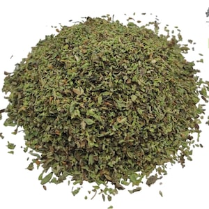 May include: A mound of dried, crushed green herbs with varying shades of olive and dark brown leaves. In the upper right corner, there is a small black tree logo with the text AGORA MARKET and a green square organic certification label.
