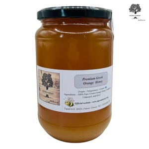 Op de afbeelding: Een pot Premium Griekse Sinaasappelhoning met een zwarte deksel. Het etiket luidt "Premium Greek Orange Honey" en vermeldt de herkomst en ingrediënten. De pot is gevuld met een goudkleurige vloeistof.