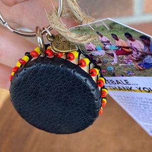 May include: A small black leather drum keychain with a yellow, red, and white beaded trim. The keychain is attached to a silver key ring. The keychain is accompanied by a tag that reads "EBALE... THANK YOU" and features a photo of children making crafts.