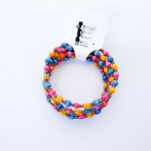 Puede incluir: Un brazalete de múltiples hilos hecho de cuentas de papel de colores. Las cuentas son en tonos de azul, rosa y amarillo. El brazalete está envuelto alrededor de una tarjeta blanca con el texto "RESTORE. UPLIFT. THRIVE. HOPE."