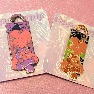 Puede incluir: Dos pines de esmalte que representan a un personaje de dibujos animados con una cola de tigre. El personaje está representado en una pose relajada, con un pin que presenta un esquema de color morado y rosa y el otro un esquema de color verde y dorado. Ambos pines tienen el texto "Happywhip" en la parte inferior.