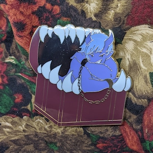 Pode incluir: Um pin de esmalte roxo com uma criatura azul semelhante a um demônio acorrentada dentro de um baú do tesouro. O baú tem uma boca grande e aberta com dentes brancos. O pin tem uma borda dourada.