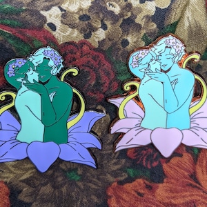 Puede incluir: Dos pines de esmalte que representan a una pareja abrazándose dentro de una flor. La pareja de la izquierda es verde y azul, mientras que la pareja de la derecha es azul y azul claro. Ambos pines tienen un tallo amarillo y una flor morada.