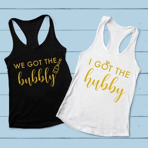 Pode incluir: Duas regatas com texto dourado em tecido preto e branco. A regata preta diz "We got the bubbly" com um gráfico de garrafa de champanhe. A regata branca diz "I got the hubby" com um gráfico de anel de diamantes.