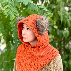 May include: A crocheted orange hooded cowl with faux fur ear flaps. The cowl is made with a single crochet stitch and has a simple design.
