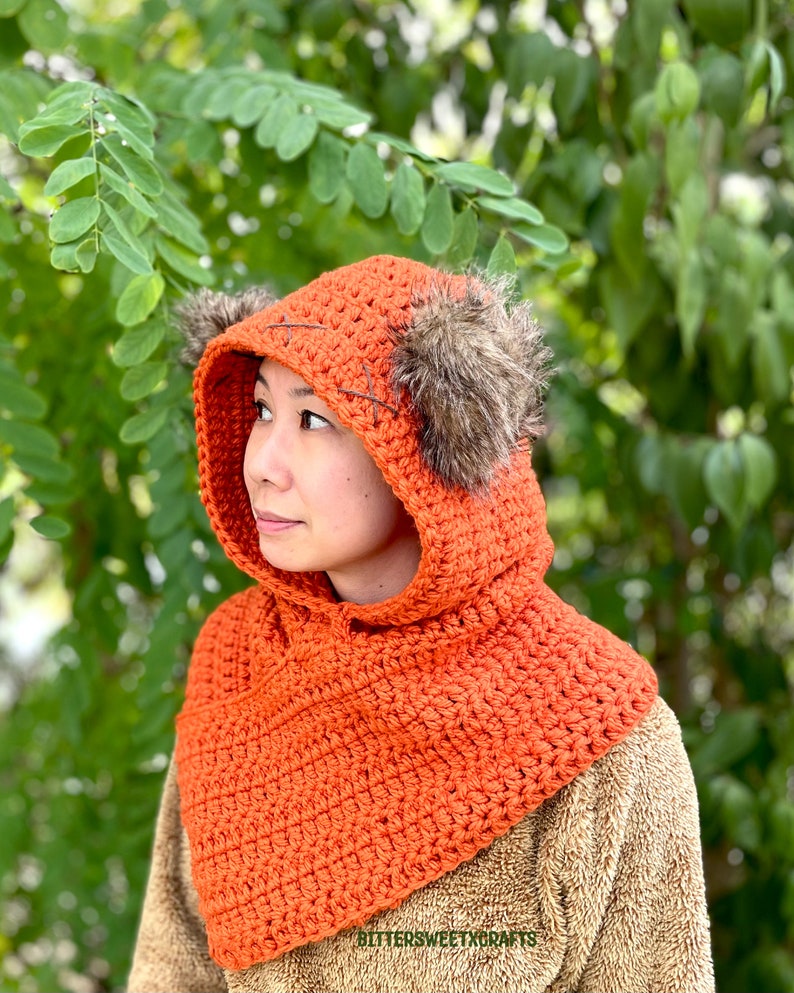May include: A crocheted orange hooded cowl with faux fur ear flaps. The cowl is made with a single crochet stitch and has a simple design.