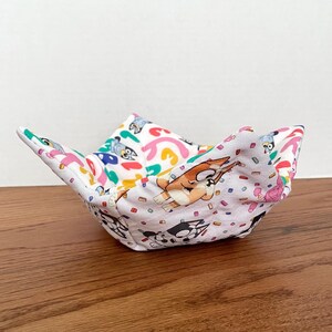 May include: A white fabric bowl with a colorful cartoon print featuring dogs and confetti. The bowl is folded and sitting on a wooden surface.