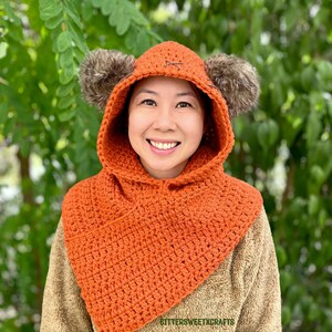 May include: A crocheted orange hooded cowl with brown fur earmuffs. The cowl has a simple stitch pattern and is worn by a person with a smile. The text "BETTERSWEETCRAFTS" is visible on the cowl.