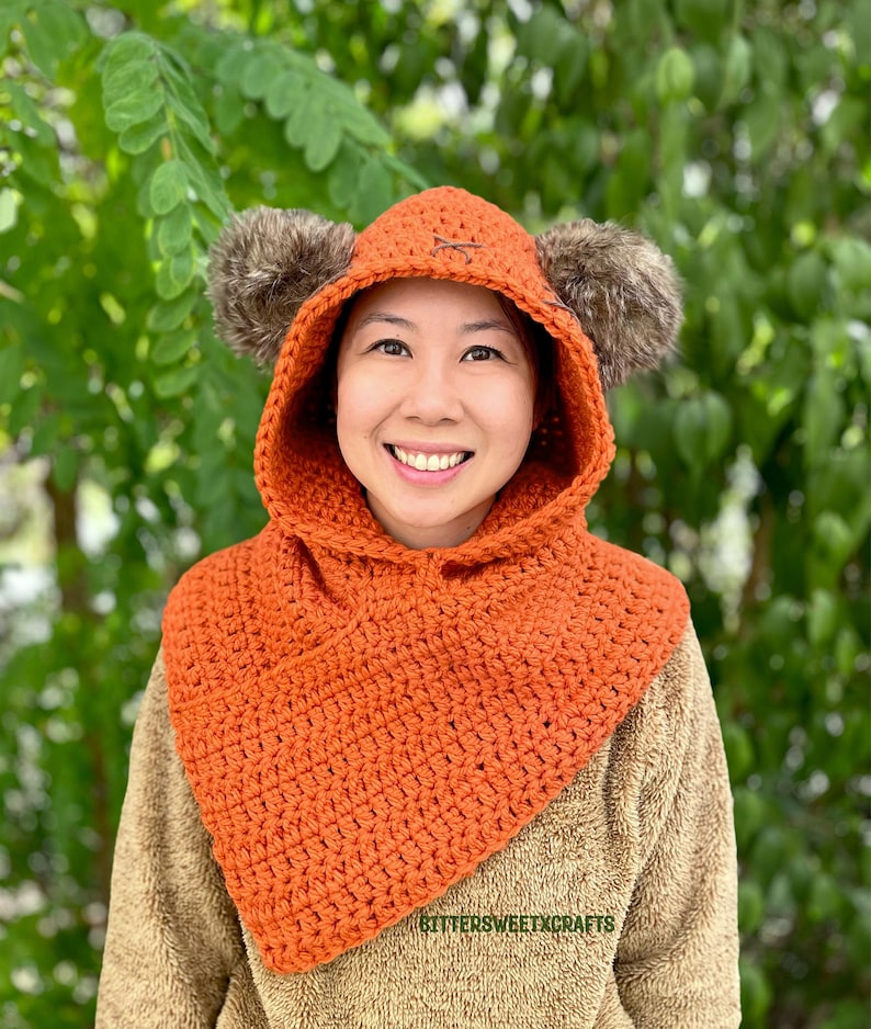 May include: A crocheted orange hooded cowl with brown fur earmuffs. The cowl has a simple stitch pattern and is worn by a person with a smile. The text "BETTERSWEETCRAFTS" is visible on the cowl.