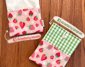 May include: Two pink fabric pouches with a strawberry print. One pouch has a white muslin lining and the other has a green gingham lining. Both pouches have a button closure.
