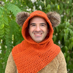 May include: A crocheted orange hooded cowl with fur earmuffs. The cowl has a pointed front and is worn by a person.