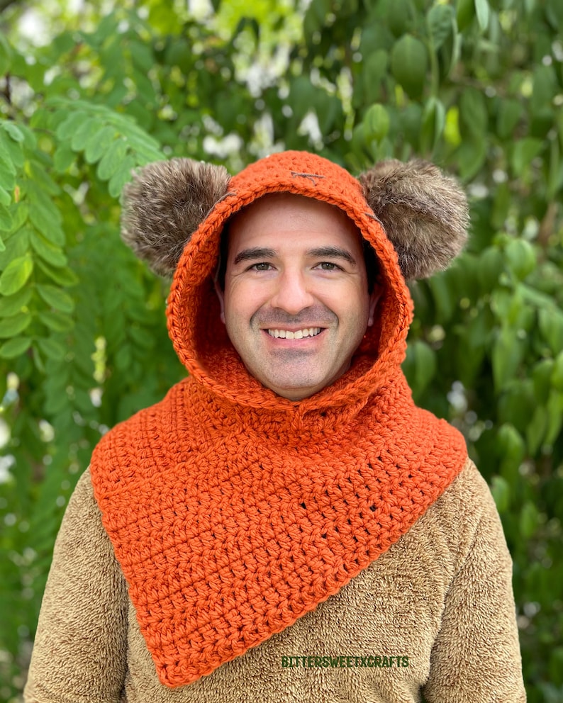 May include: A crocheted orange hooded cowl with fur earmuffs. The cowl has a pointed front and is worn by a person.