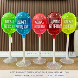 May include: Five clear plastic lollipop holders with white sticks and bases. The lollipop holders are in the colours green, light blue, pink, red, and yellow. Each lollipop holder has a white label with the text "ARIADNA'S 3RD BIRTHDAY" printed on it.
