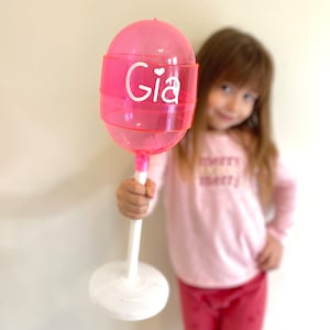 May include: A pink plastic lollipop with a white base and a white sticker that says "Gia" with a small heart above the name. The lollipop is being held by a person.