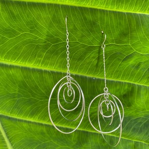 May include: Silver dangle earrings with multiple interlocking circles. The earrings are hanging against a green leaf background.