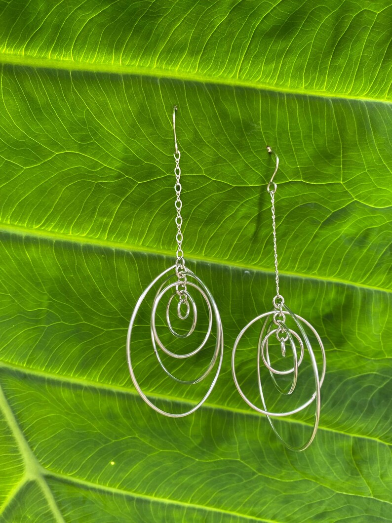 May include: Silver dangle earrings with multiple interlocking circles. The earrings are hanging against a green leaf background.
