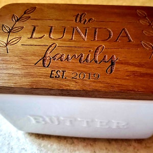 May include: A white ceramic butter dish with a wooden lid. The lid is engraved with the text "the LUNDA family EST. 2019" and features a leafy design.