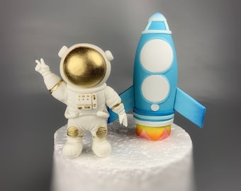 以下が含まれることがあります： 金色のヘルメットをかぶった白い宇宙飛行士のフィギュアと、青と白のロケットのフィギュア。宇宙飛行士は片手をピースサインにして掲げている。ロケットは青とオレンジの炎を出している。