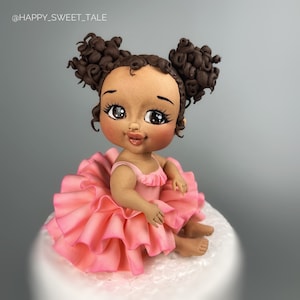 Könnte beinhalten: Eine braungefärbte Baby-Puppe mit braunen lockigen Haaren in Zöpfen, die ein rosa Rüschenkleid trägt. Die Puppe sitzt auf einem weißen Kuchen.