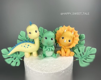 May include: Three adorable dinosaur cake toppers, a yellow dinosaur, a green dinosaur, and a yellow triceratops, with green leaves on a white cake.
