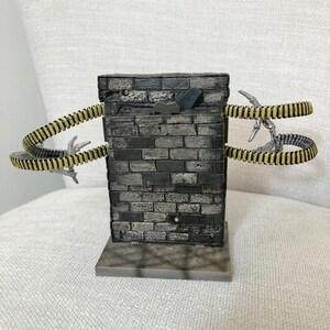 May include: A detailed miniature diorama featuring a brick wall structure with two segmented, tentacle-like appendages extending outwards. The tentacles are yellow and black, with silver claw-like tips. The wall is gray and black.