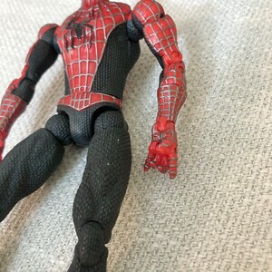 May include: A Spider-Man action figure with a red and black costume. The figure has a detailed design with a web pattern on the red sections. The figure's joints are visible, and the hands are in a gripping pose.