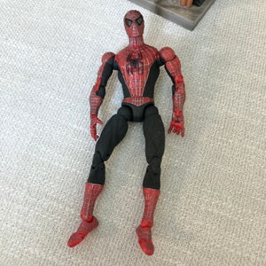May include: A Spider-Man action figure, featuring a red and black costume with a spider emblem on the chest. The figure has articulated joints and detailed paintwork, showcasing the character's iconic design. The figure is approximately 6 inches tall.