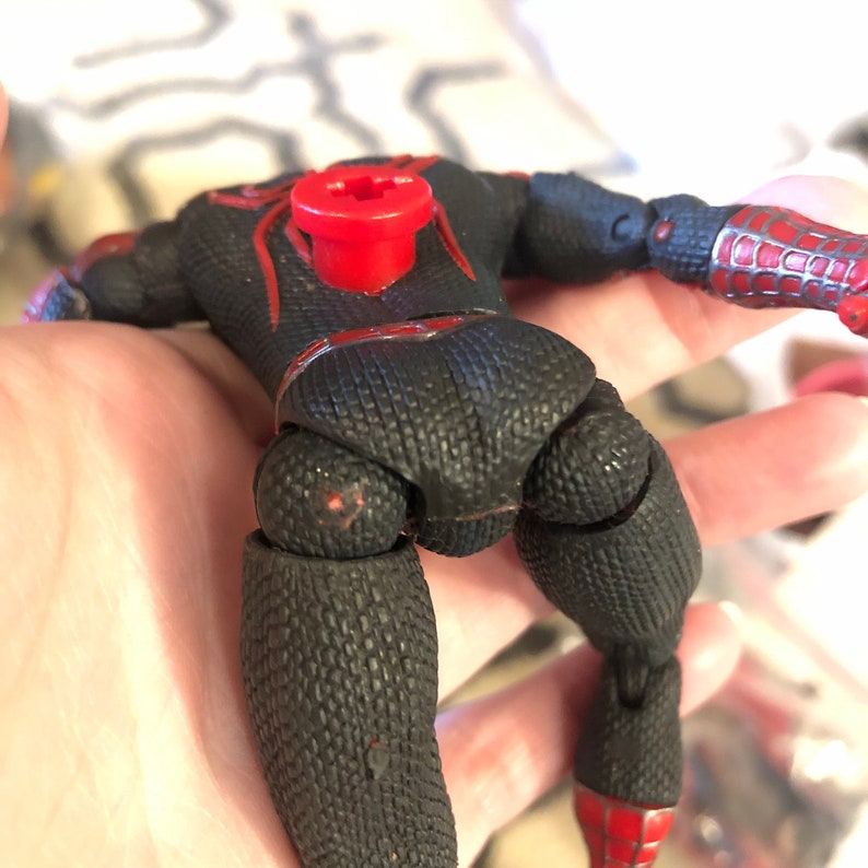 May include: Black and red action figure of a superhero, featuring a textured black suit with red accents. The figure has articulated joints and a red spider emblem on the back. The figure's arms and legs are visible.
