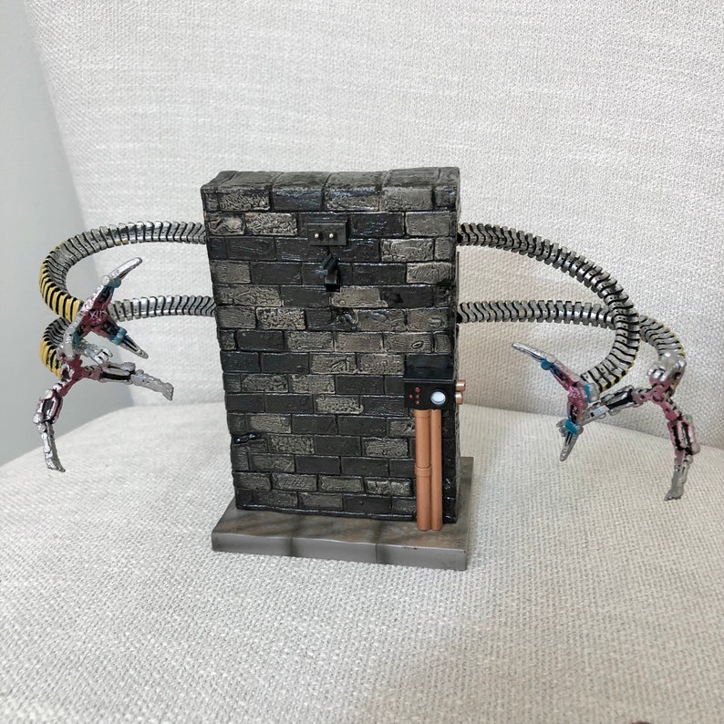 May include: A detailed sculpture of a brick structure with four metallic, tentacle-like arms extending outwards. The brickwork is dark gray, and the arms are silver with red, yellow, and blue accents. The base is gray.