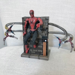 May include: A Spider-Man action figure in a red and black suit, posed against a gray brick wall. The figure is attached to mechanical arms with metallic silver and red accents. The base is gray.
