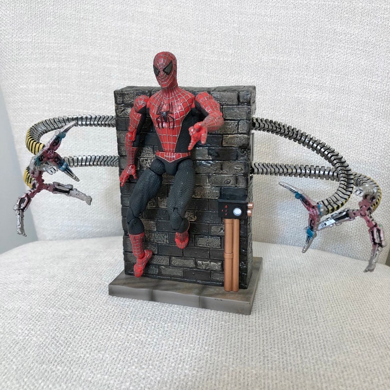 May include: A Spider-Man action figure in a red and black suit, posed against a gray brick wall. The figure is attached to mechanical arms with metallic silver and red accents. The base is gray.
