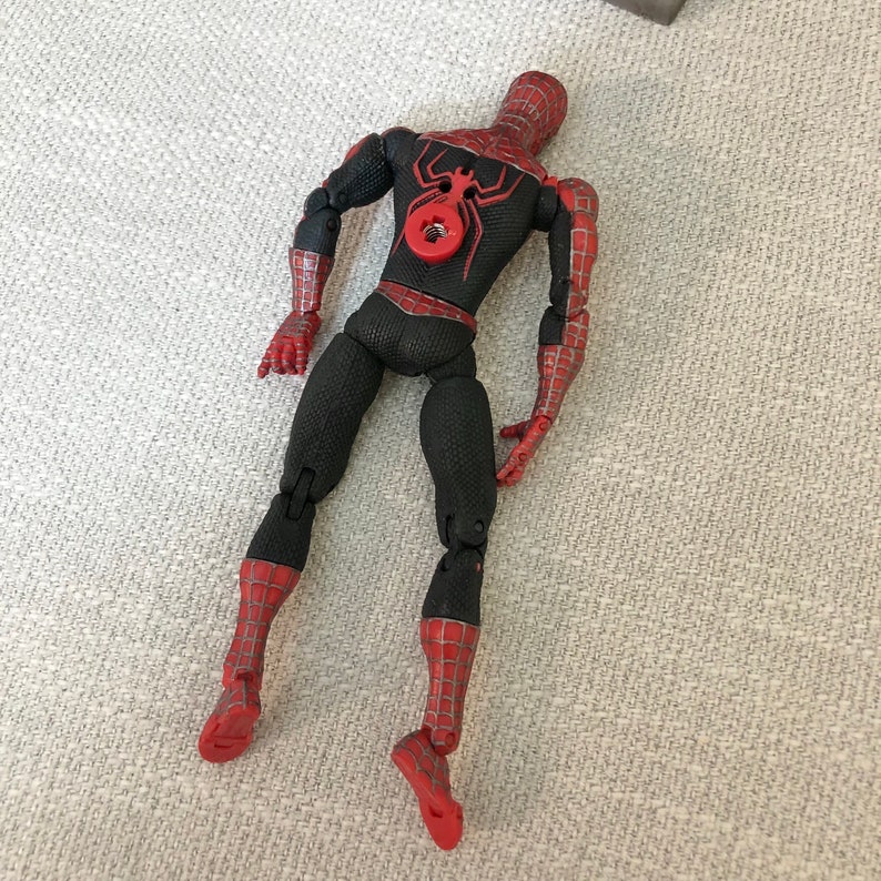 May include: A Spider-Man action figure in a crouching pose. The figure is primarily black with red accents, including the hands, feet, and sections of the arms and legs. A red spider emblem is on the back. The figure is lying on a textured, light gray surface.