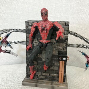 May include: A Spider-Man action figure in a red and black suit, posed against a gray brick wall. The figure has a detailed design and is reaching out. Metallic tentacles are visible in the background, suggesting a battle scene.