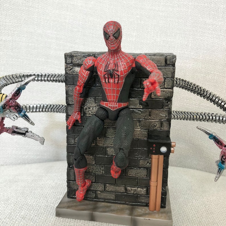 May include: A Spider-Man action figure in a red and black suit, posed against a gray brick wall. The figure has a detailed design and is reaching out. Metallic tentacles are visible in the background, suggesting a battle scene.