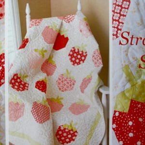 May include: A white quilt with a patchwork design featuring red and pink strawberries. The quilt is draped over a white chair with a green and white patterned fabric behind it. The text "Strawberry Shortcake" is visible in the background.