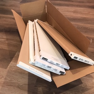 May include: A cardboard box filled with several white window blinds. The blinds are packaged in white and brown cardboard. The box is open, revealing the contents inside. The blinds are stacked neatly.
