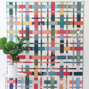 May include: A colorful quilt with a plaid design, featuring intersecting stripes in red, blue, green, orange, and yellow. The quilt is displayed on a white wall. Text on the quilt reads "Plaidy Cake". The quilt is a layer cake or fat quarter quilt in 5 sizes.