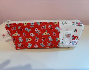 May include: A rectangular pencil case with a zipper closure. The case is made of fabric with a red floral pattern on one side and a white pattern with houses and trees on the other. The label reads "MAYHEWS FABRIC".