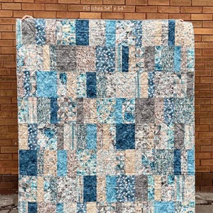 May include: A patchwork quilt with a variety of blue, gray, and tan fabrics. The quilt is rectangular and measures 54" x 64". The text "SLICED BREAD" is at the top, with "MY BESTE QUILT CO." and a pattern description at the bottom.