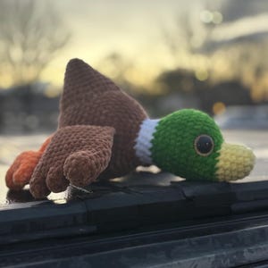May include: A handmade crochet duck toy. The duck features a brown body, a green head with a white neck ring, and a yellow beak. The duck's feet are orange. The toy is lying down on a dark surface.