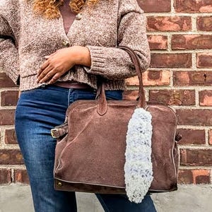 May include: A brown suede tote bag with a matching handle and a decorative white and gray fluffy charm. The bag has a rectangular shape with a buckle detail and is held in front of a brick wall.