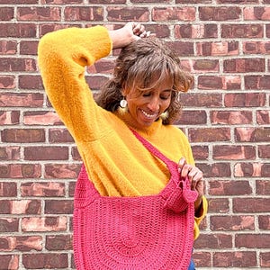 May include: A bright pink, crocheted shoulder bag with a rounded shape. The bag is held by a person wearing a yellow sweater and blue jeans. The background is a brick wall.