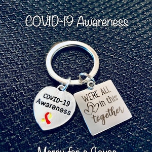 May include: Silver keychain with two charms. One charm is a heart with the text "COVID-19 Awareness" and a red ribbon. The other charm is a square with the text "WE'RE ALL in this together" and two hearts. The text "Merry for a Cause" is below the charms.