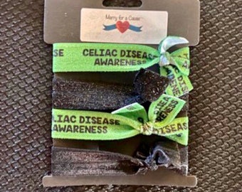 May include: A set of four hair ties with green and black bands. The bands have the text "Celiac Disease Awareness" printed on them. The hair ties are packaged on a card with the text "Merry for a Cause".
